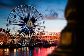 A large Ferris wheel with a prominent Mickey Mouse face in the center, illuminated against the evening sky. The structure is lit up with various colors, and there are visible roller coasters and other amusement park rides in the background, with their reflection on the water below.