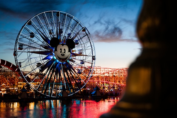 A large Ferris wheel with a prominent Mickey Mouse face in the center, illuminated against the evening sky. The structure is lit up with various colors, and there are visible roller coasters and other amusement park rides in the background, with their reflection on the water below.