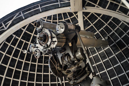A large telescope is situated inside an observatory dome with a grid-like supporting structure. The telescope is composed of metallic components, intricate gears, and appears to be designed for astronomical observations.