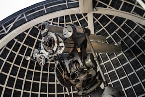 A large telescope is situated inside an observatory dome with a grid-like supporting structure. The telescope is composed of metallic components, intricate gears, and appears to be designed for astronomical observations.