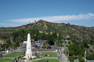 A scenic landscape featuring a large, tall monument in a park with people around it. In the background, there is a hilly terrain covered with green vegetation under a clear blue sky. Several cars are parked in a lot behind the monument, and the overall setting appears to be a popular outdoor destination with numerous visitors.