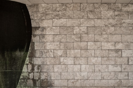 The image features a textured stone wall with rectangular stone tiles arranged in a grid pattern. On the left side, a dark, curved area interrupts the uniformity of the tiles, possibly indicating water damage or an artistic design element.