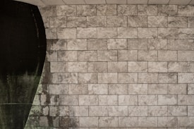 The image features a textured stone wall with rectangular stone tiles arranged in a grid pattern. On the left side, a dark, curved area interrupts the uniformity of the tiles, possibly indicating water damage or an artistic design element.