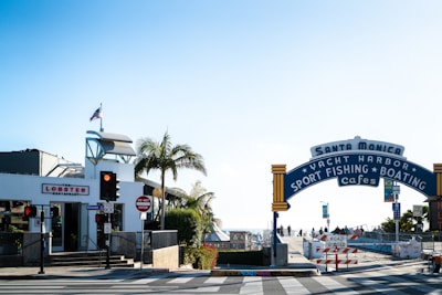A sunny street view featuring the iconic Santa Monica Yacht Harbor sign, with palm trees and a lobster restaurant to the left. The intersection includes traffic lights and signs, along with some pedestrians in the background.