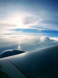 An airplane wing appears above a layer of fluffy white clouds with a serene blue sky overhead. Sunlight creates a soft glow across the clouds, adding a peaceful atmosphere to the scene.