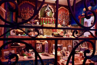 An intricately decorated altar featuring a variety of elements including skull motifs, religious icons, vibrant papel picado, candles, and offerings like fruits. Photographs and personal mementos are arranged on multiple levels, surrounded by bright colors and festive decorations.