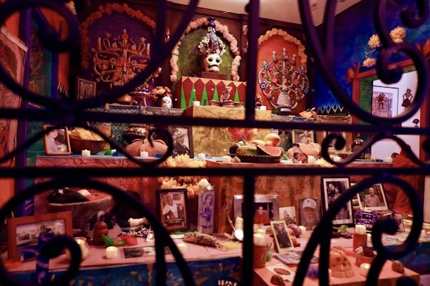 An intricately decorated altar featuring a variety of elements including skull motifs, religious icons, vibrant papel picado, candles, and offerings like fruits. Photographs and personal mementos are arranged on multiple levels, surrounded by bright colors and festive decorations.