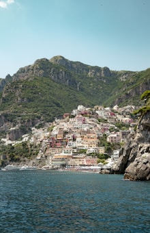A picturesque coastal town with colorful buildings stacked on steep hillsides, surrounded by lush green cliffs. The clear blue waters of the sea contrast beautifully with the vibrant architecture and the surrounding natural landscape.