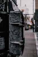 A cartoon-like character is drawn on a weathered, metal surface of an urban object. The background features a blurred street with a hint of buildings and soft lights.