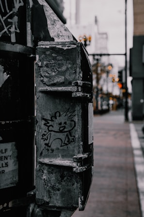 A cartoon-like character is drawn on a weathered, metal surface of an urban object. The background features a blurred street with a hint of buildings and soft lights.