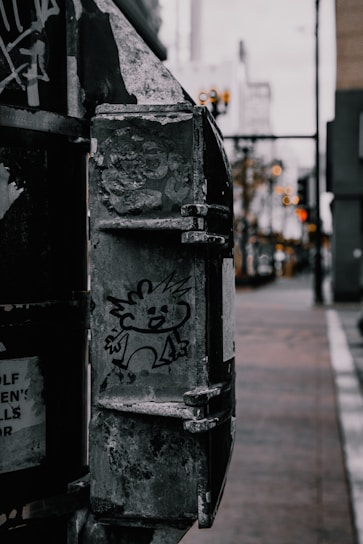 A cartoon-like character is drawn on a weathered, metal surface of an urban object. The background features a blurred street with a hint of buildings and soft lights.