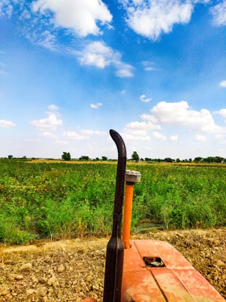 A vast agricultural field stretches under a bright blue sky filled with scattered white clouds. In the foreground, the top of a tractor is visible, surrounded by green crops and dry soil.