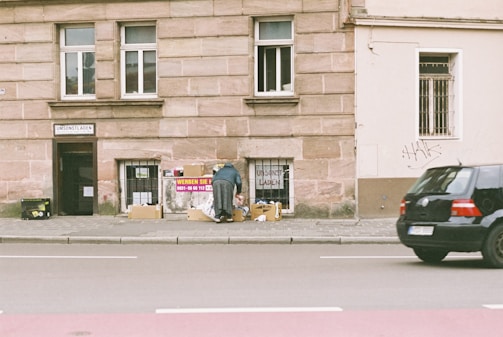 A person is bending over to look through boxes placed on the sidewalk in front of a building with a sign that reads 'UMSONSTLADEN.' The wall is made of bricks and has windows with bars. Some graffiti is present on the wall, and a black car is parked on the street next to the boxes.