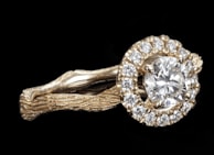 A gold ring features a large round diamond at its center, surrounded by a halo of smaller diamonds. The band of the ring has a textured, organic appearance, resembling intertwined branches or vines, lending a unique and intricate design.