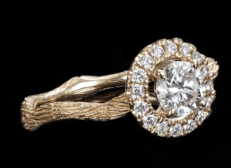A gold ring features a large round diamond at its center, surrounded by a halo of smaller diamonds. The band of the ring has a textured, organic appearance, resembling intertwined branches or vines, lending a unique and intricate design.