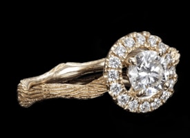 A gold ring features a large round diamond at its center, surrounded by a halo of smaller diamonds. The band of the ring has a textured, organic appearance, resembling intertwined branches or vines, lending a unique and intricate design.