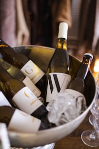 A chilled silver bowl contains multiple bottles of beverages, including wine and ginger ale, surrounded by ice cubes. The labels on the bottles are prominently displayed, featuring bold letters and minimalistic designs. The setting gives a sophisticated atmosphere, suggesting a gathering or event.
