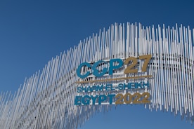 A contemporary art installation with vertical metal rods arranged in a wave pattern forms a striking visual backdrop. The metal rods create a textured appearance against the clear blue sky. Prominent text reads 'COP27 SHARM EL-SHEIKH EGYPT 2022' in bold letters, alternating between blue and gold colors, signifying a significant event.