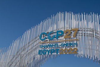 A contemporary art installation with vertical metal rods arranged in a wave pattern forms a striking visual backdrop. The metal rods create a textured appearance against the clear blue sky. Prominent text reads 'COP27 SHARM EL-SHEIKH EGYPT 2022' in bold letters, alternating between blue and gold colors, signifying a significant event.