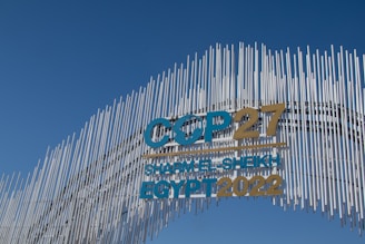A contemporary art installation with vertical metal rods arranged in a wave pattern forms a striking visual backdrop. The metal rods create a textured appearance against the clear blue sky. Prominent text reads 'COP27 SHARM EL-SHEIKH EGYPT 2022' in bold letters, alternating between blue and gold colors, signifying a significant event.