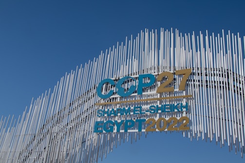 A contemporary art installation with vertical metal rods arranged in a wave pattern forms a striking visual backdrop. The metal rods create a textured appearance against the clear blue sky. Prominent text reads 'COP27 SHARM EL-SHEIKH EGYPT 2022' in bold letters, alternating between blue and gold colors, signifying a significant event.