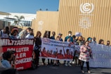 A diverse group of people are gathered outside a building with climate-related insignia. They are holding various banners with messages supporting human rights, indigenous rights, and anti-militarism. One banner reads 'DEFEND HUMAN RIGHTS AND THE RIGHTS OF INDIGENOUS PEOPLE,' and another reads 'MILITARISM POLLUTER COLONIZER.' A photographer is taking pictures of the group.