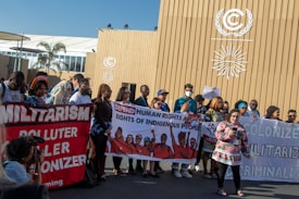 A diverse group of people are gathered outside a building with climate-related insignia. They are holding various banners with messages supporting human rights, indigenous rights, and anti-militarism. One banner reads 'DEFEND HUMAN RIGHTS AND THE RIGHTS OF INDIGENOUS PEOPLE,' and another reads 'MILITARISM POLLUTER COLONIZER.' A photographer is taking pictures of the group.