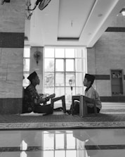 Two children are seated on the floor facing each other in a spacious room with large windows. Both are wearing traditional caps and are engaged in reading or recitation, supported by small wooden lecterns. The room has a reflective tiled floor, a ceiling fan, and a bookcase in the background.