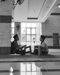 Two children are seated on the floor facing each other in a spacious room with large windows. Both are wearing traditional caps and are engaged in reading or recitation, supported by small wooden lecterns. The room has a reflective tiled floor, a ceiling fan, and a bookcase in the background.