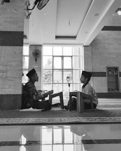 Two children are seated on the floor facing each other in a spacious room with large windows. Both are wearing traditional caps and are engaged in reading or recitation, supported by small wooden lecterns. The room has a reflective tiled floor, a ceiling fan, and a bookcase in the background.