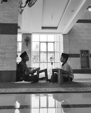Two children are seated on the floor facing each other in a spacious room with large windows. Both are wearing traditional caps and are engaged in reading or recitation, supported by small wooden lecterns. The room has a reflective tiled floor, a ceiling fan, and a bookcase in the background.
