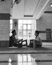 Two children are seated on the floor facing each other in a spacious room with large windows. Both are wearing traditional caps and are engaged in reading or recitation, supported by small wooden lecterns. The room has a reflective tiled floor, a ceiling fan, and a bookcase in the background.