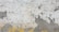 A weathered concrete wall with peeling paint exhibiting various shades of grey and white. The surface shows textures and patterns where the paint has cracked and flaked off, revealing the underlying material. A line of yellow paint runs along the bottom edge, adding contrast to the otherwise neutral tones.