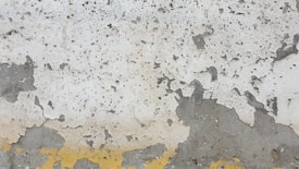 A weathered concrete wall with peeling paint exhibiting various shades of grey and white. The surface shows textures and patterns where the paint has cracked and flaked off, revealing the underlying material. A line of yellow paint runs along the bottom edge, adding contrast to the otherwise neutral tones.