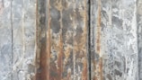 A weathered and rusted metal surface with vertical grooves and varying textures. The surface shows signs of decay with a mix of gray, brown, and orange hues.