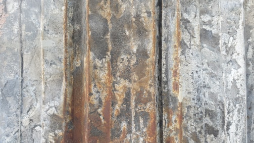 A weathered and rusted metal surface with vertical grooves and varying textures. The surface shows signs of decay with a mix of gray, brown, and orange hues.
