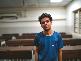A person with curly hair is standing in a classroom setting. The room has rows of empty wooden desks and a window with multiple panes. The walls are painted white and beige, and the ceiling has fluorescent lighting.