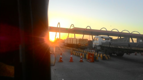A truck approaches a toll booth during sunset, with the sun casting a warm glow over the scene. The booth features several arches with red and green lights above the lanes. Traffic cones and barriers guide vehicles through the lanes.