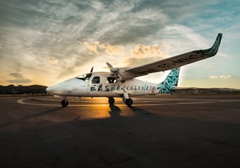 A small white airplane is parked on a runway with the text 'EAS Barcelona' on its side. The plane has distinctive teal patterns on its tail fin and wing tips. The scene is set at sunset, casting a warm glow over the aircraft with a dramatic sky filled with wispy clouds.