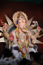 A large, intricately designed statue of Lord Ganesha, adorned with colorful garlands and elaborate jewelry. The statue features a prominent headdress with intricate details and multiple arms holding various symbolic items. The background includes decorative elements that enhance the spiritual ambiance.