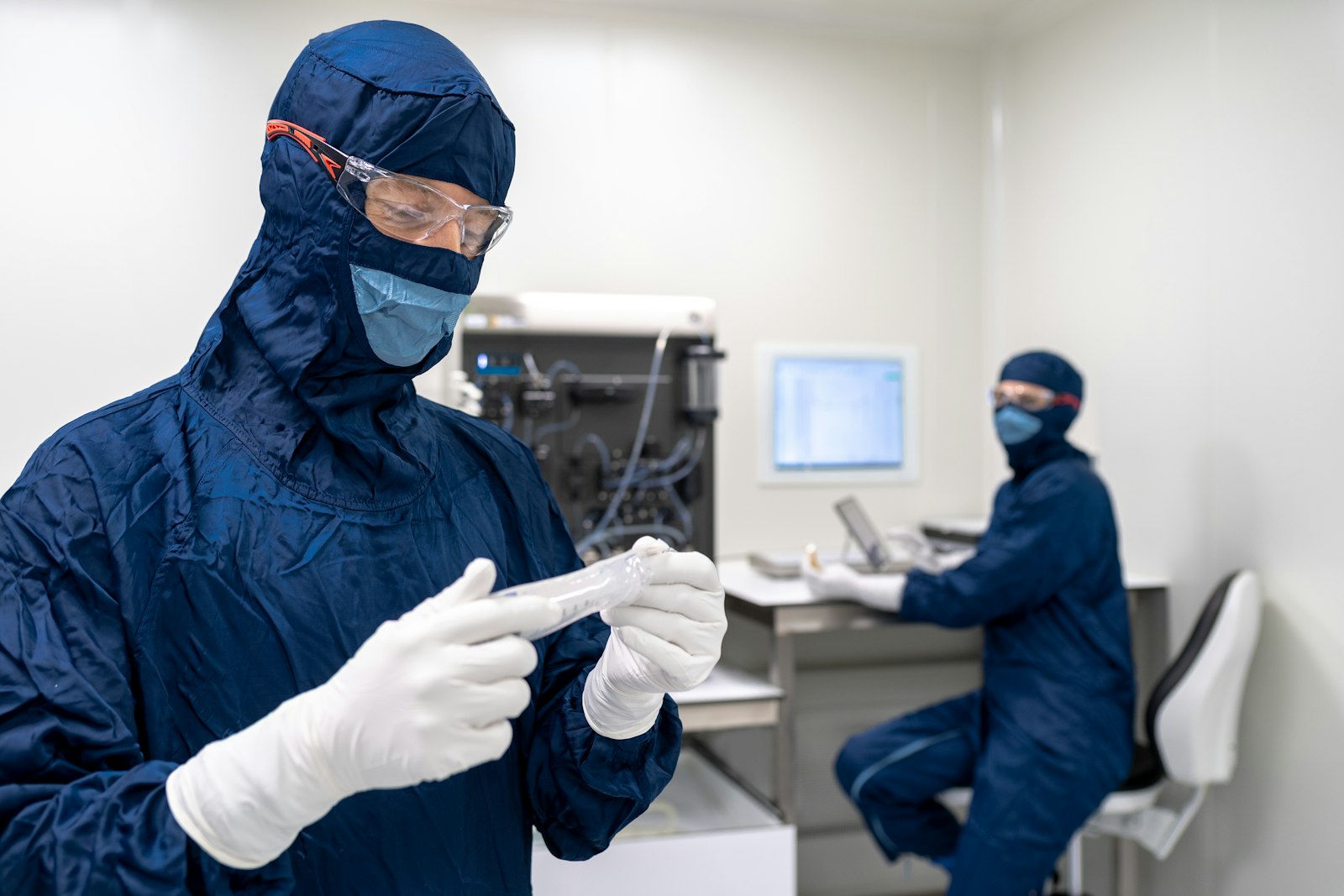 Two technicians in blue protective coveralls, gloves and face masks working side by side in a clean manufacturing room.