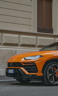 An orange luxury SUV is parked on a street against the backdrop of a beige building with textured walls and wooden shutters. The car, with its sleek lines and sporty design, has a prominent emblem on the front.