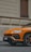 An orange luxury SUV is parked on a street against the backdrop of a beige building with textured walls and wooden shutters. The car, with its sleek lines and sporty design, has a prominent emblem on the front.