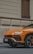 An orange luxury SUV is parked on a street against the backdrop of a beige building with textured walls and wooden shutters. The car, with its sleek lines and sporty design, has a prominent emblem on the front.