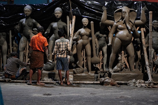 Several people work on large clay sculptures depicting figures from Hindu mythology, with an emphasis on detailed craftsmanship. The scene takes place in an artisan workshop, indicating a cultural and religious context. The figures are at various stages of completion, surrounded by tools and materials.