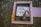 A worn wooden frame lies on a textured surface, partially surrounding an arrangement including heart-shaped sunglasses and a collage of words like 'Lights... Camera...' and 'Art'. An orange fabric piece adorns the edge of the frame, adding a splash of color.