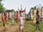 A group of colorful, stylized metal sculptures resembling human figures is arranged outdoors on a grassy area. The sculptures are of varying colors including red, blue, yellow, and earth tones. Each sculpture has unique features, such as horns or distinct facial expressions, and they appear to be made from recycled materials. Trees and foliage form the background, creating a natural setting for the artwork.