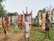 A group of colorful, stylized metal sculptures resembling human figures is arranged outdoors on a grassy area. The sculptures are of varying colors including red, blue, yellow, and earth tones. Each sculpture has unique features, such as horns or distinct facial expressions, and they appear to be made from recycled materials. Trees and foliage form the background, creating a natural setting for the artwork.