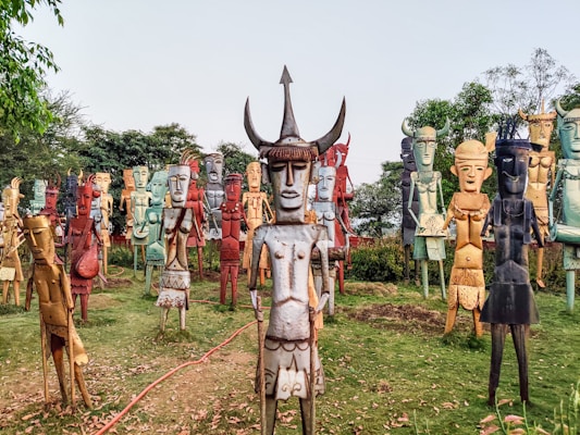 A group of colorful, stylized metal sculptures resembling human figures is arranged outdoors on a grassy area. The sculptures are of varying colors including red, blue, yellow, and earth tones. Each sculpture has unique features, such as horns or distinct facial expressions, and they appear to be made from recycled materials. Trees and foliage form the background, creating a natural setting for the artwork.