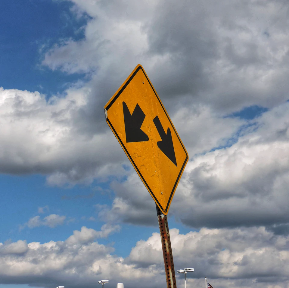 Yellow diamond-shaped road sign mounted on a metal pole under blue sky with arrows pointing